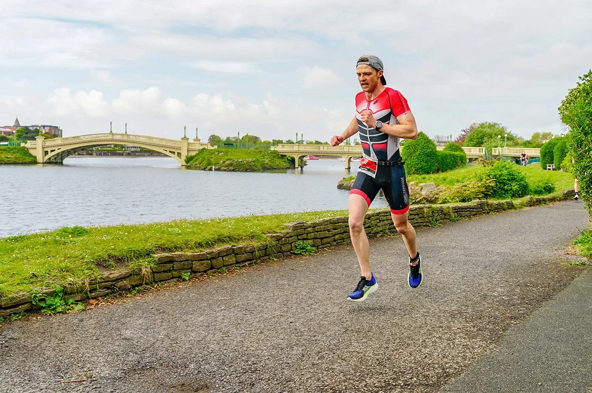 A man in a red and black triathlon suit runs along a riverside path with a bridge in the background during a race.