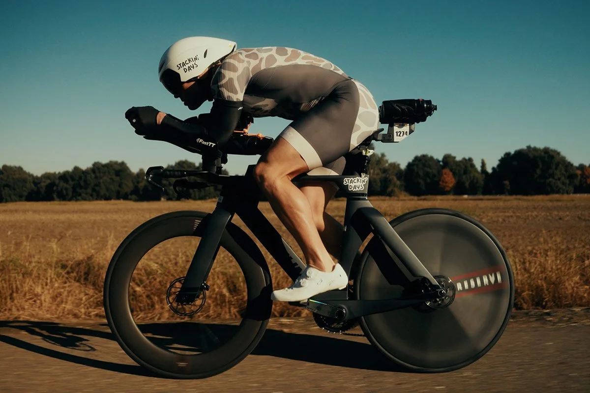 A cyclist in an aerodynamic position rides a time-trial bike along a rural road, wearing a helmet and streamlined racing gear.