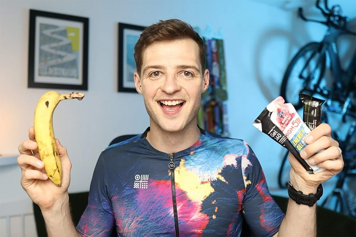 Smiling man in a colorful cycling jersey holding a banana and energy gel packets indoors.