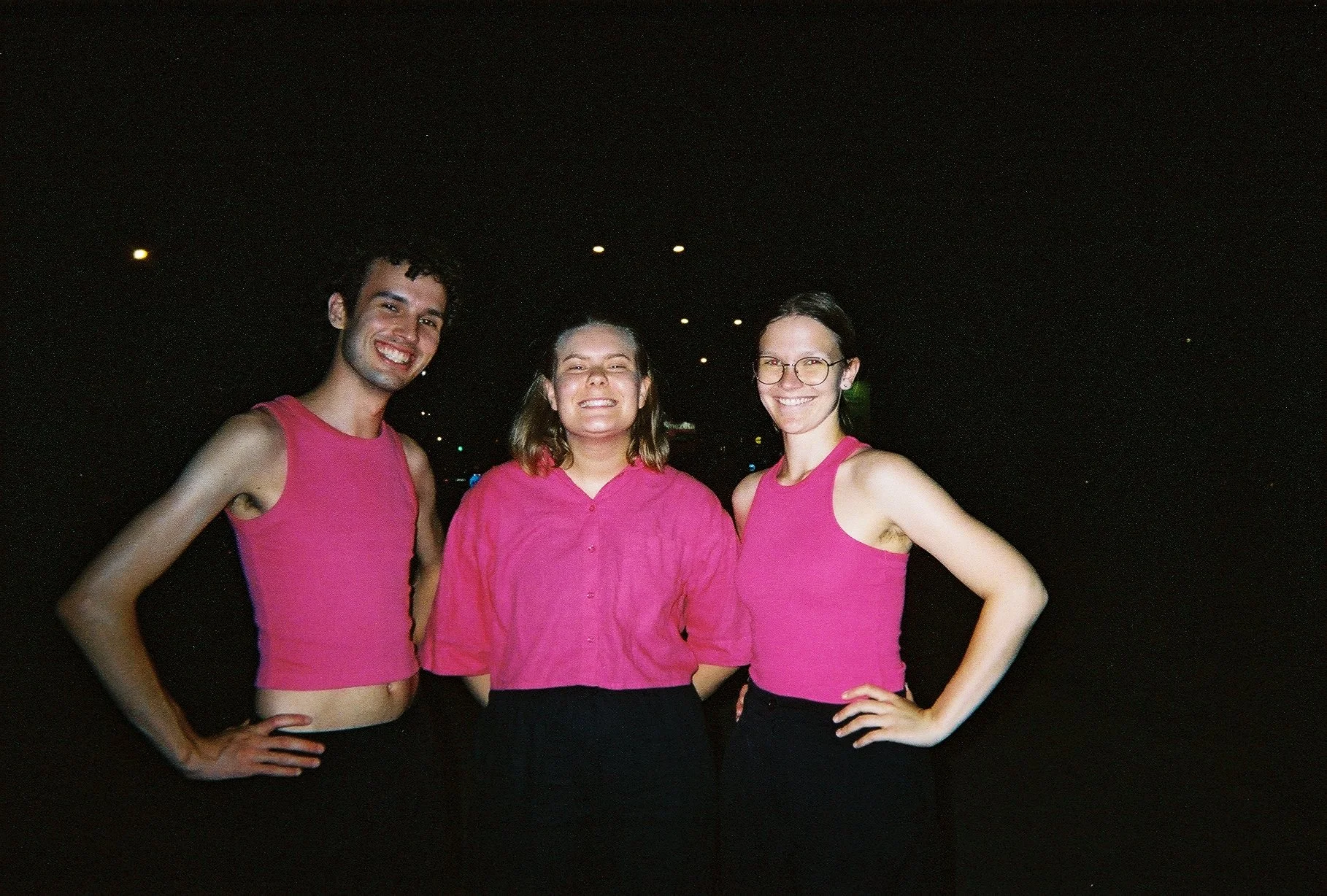 Tre personer smiler og poserer for kameraet, to kvinner og en mann, i mørket. Alle er iført rosa klær.