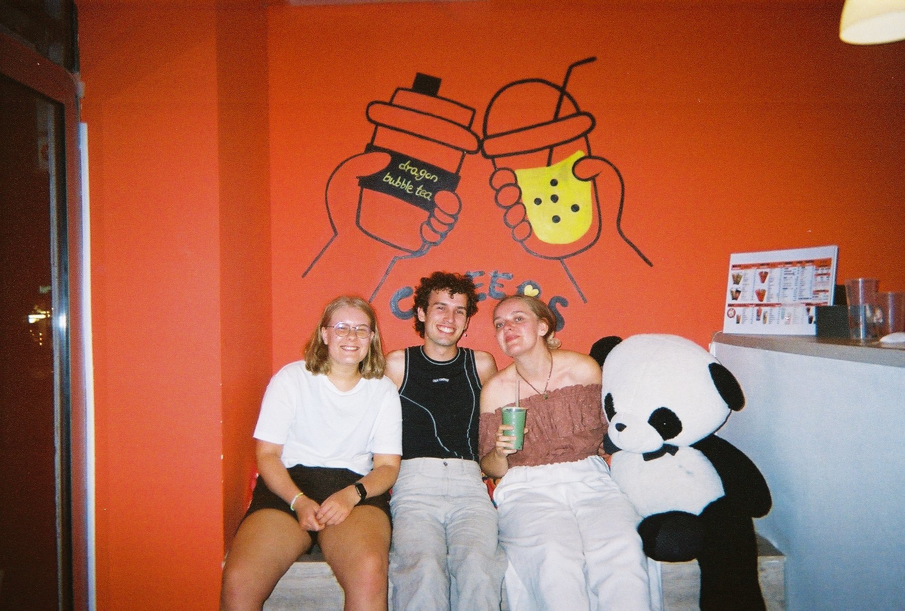 Tre mennesker sitter sammen og smiler foran en vegg med en hobby-illustrasjon av to hender som holder koppene sine, mens de to koppene har tekopper med bubble tea. En av personene holder en grønn kopp, og foran dem er en stor pandaskulptur. Det er en
