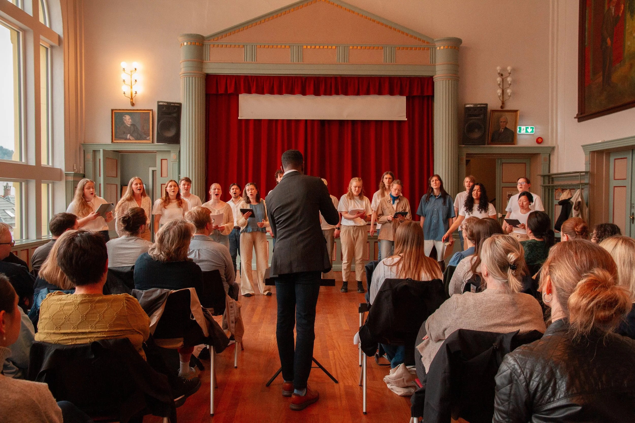 En gruppe personer står på scenen og synger, dirigent dirigerer, publikummet sitter og ser på innendørs, med store vinduer på venstre side og røde gardiner.