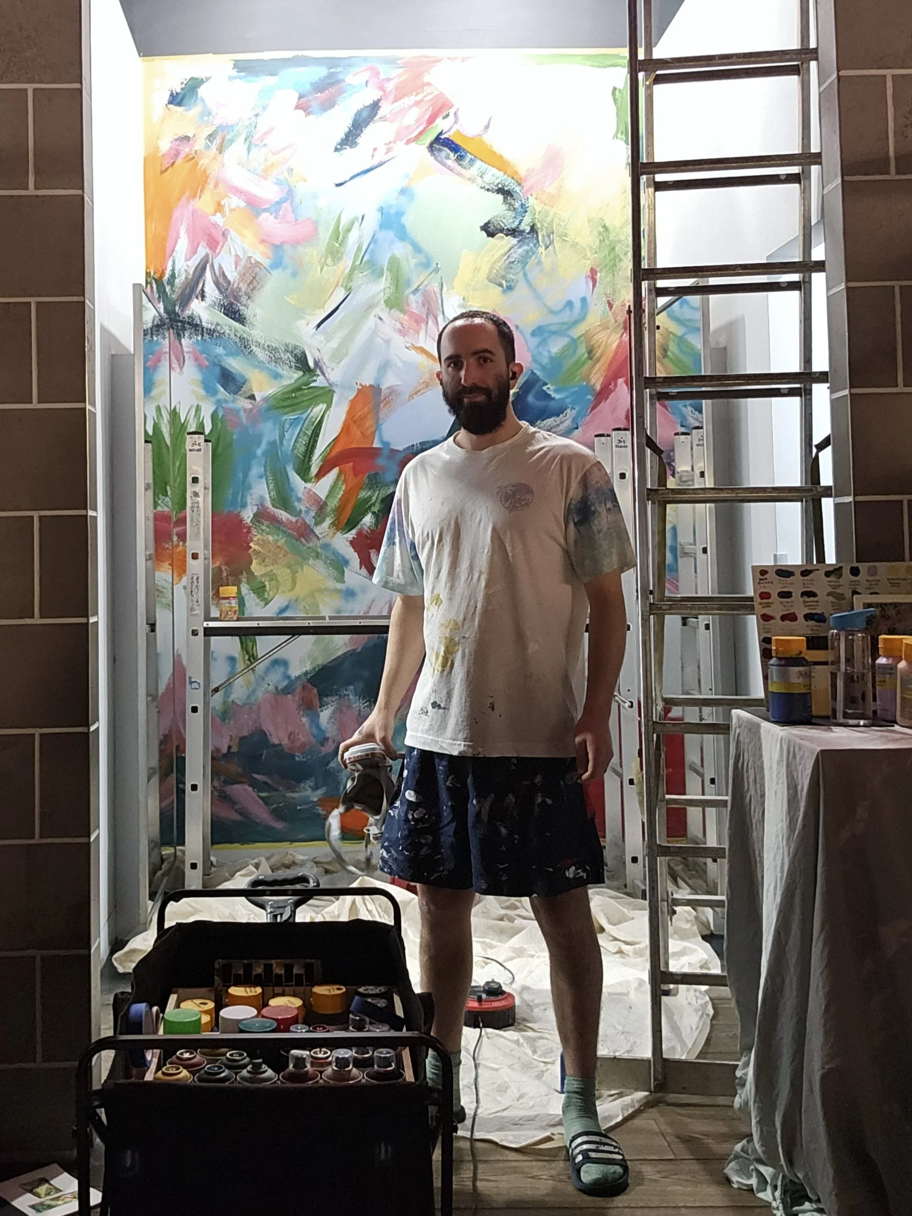 A man with a beard and tattoo on his arm, wearing a white t-shirt, shorts, slippers, and socks, stands in front of a colorful abstract painting in an art studio. There is a ladder on the right, and various art supplies on a table and in a container in the foreground.
