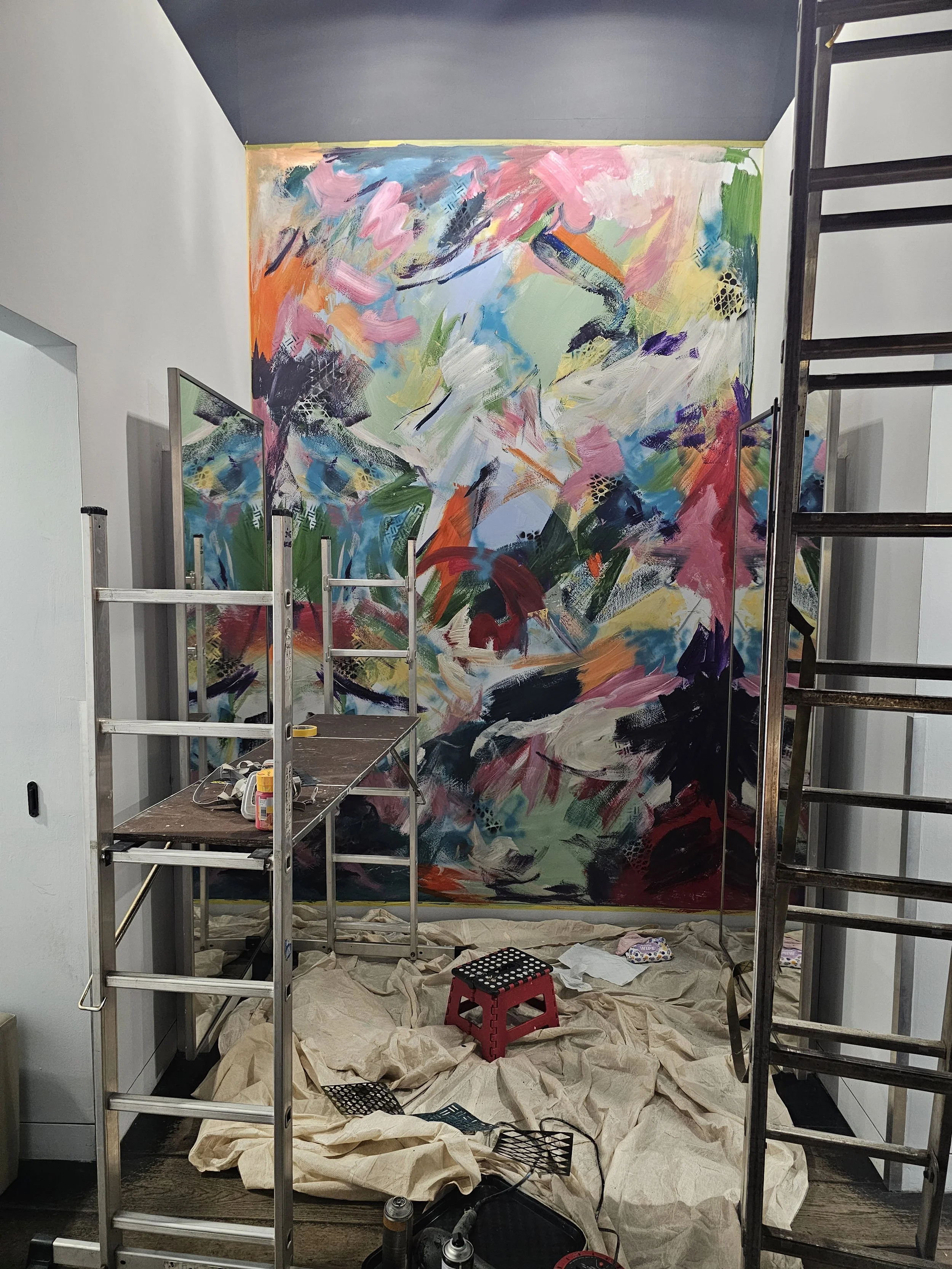 Mural installation in progress - colourful abstract painting on large wall at Selfridges Birmingham, surrounded by scaffolding, ladders and art supplies.