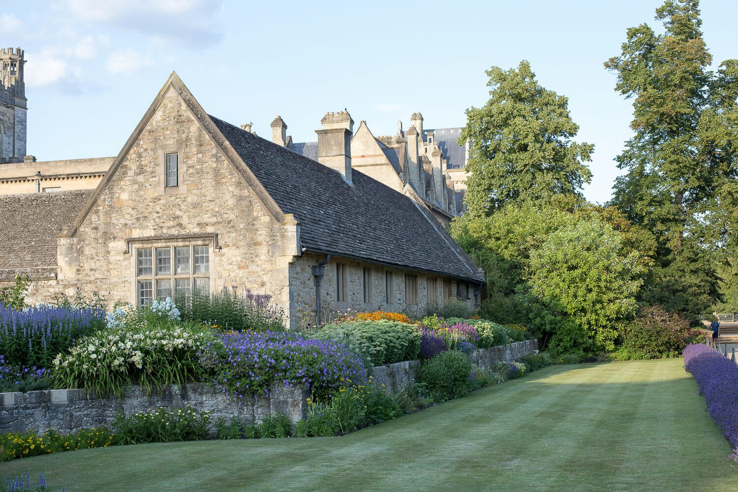 Stone cottage with a dark shingled roof, surrounding colorful flower garden, tall trees, and a well-maintained lawn under a blue sky.