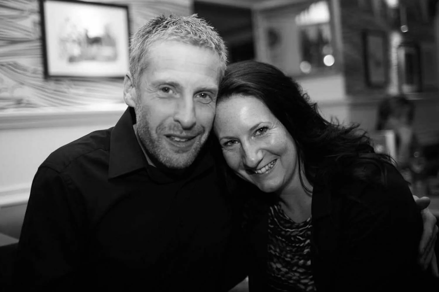 A black and white photo of Phil and Danni sitting together in a casual setting, with artwork on the walls.
