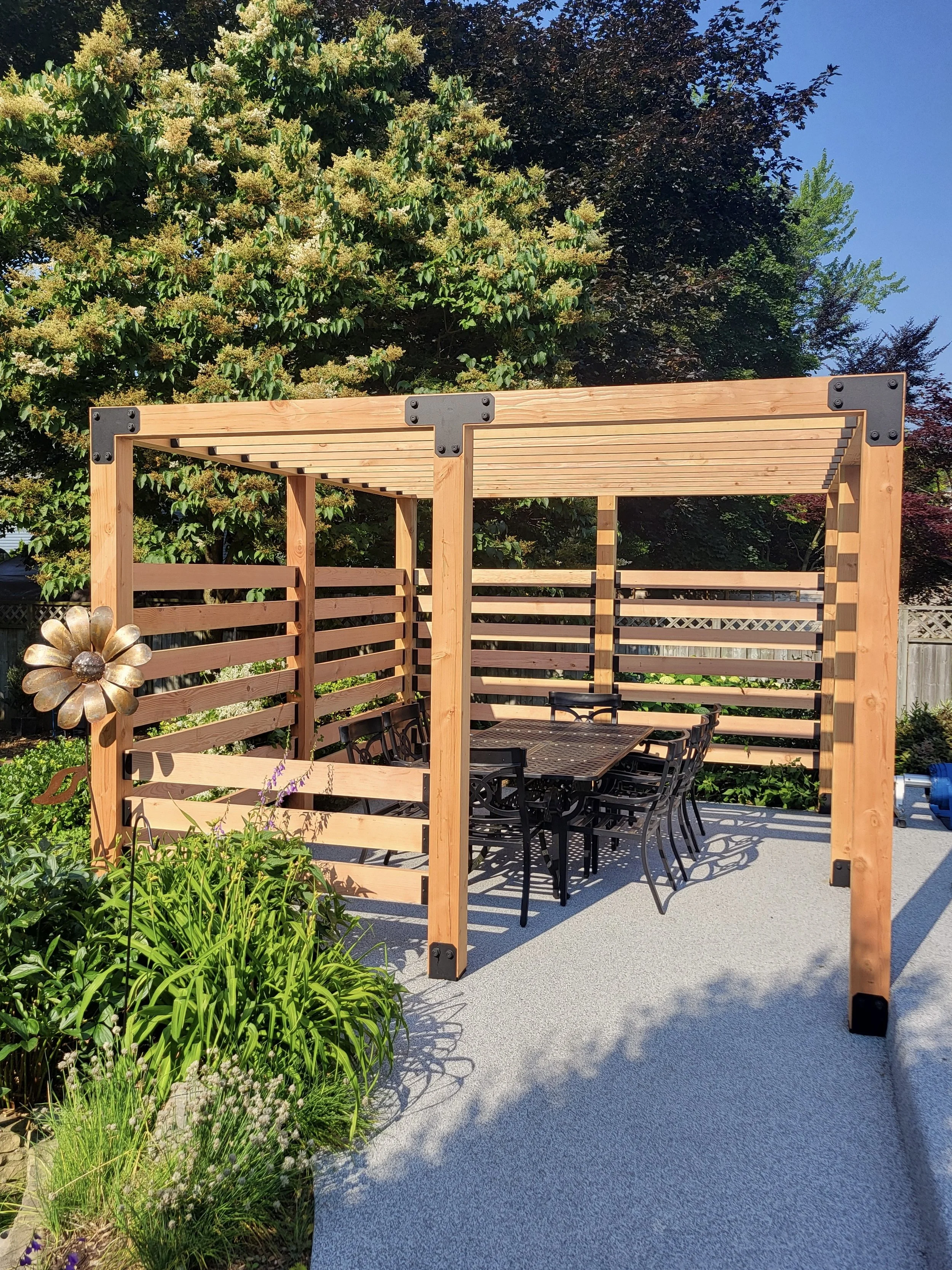 A wooden outdoor pergola with a black metal dining table and chairs underneath, situated on a concrete patio, surrounded by green plants and trees with a bright blue sky overhead.