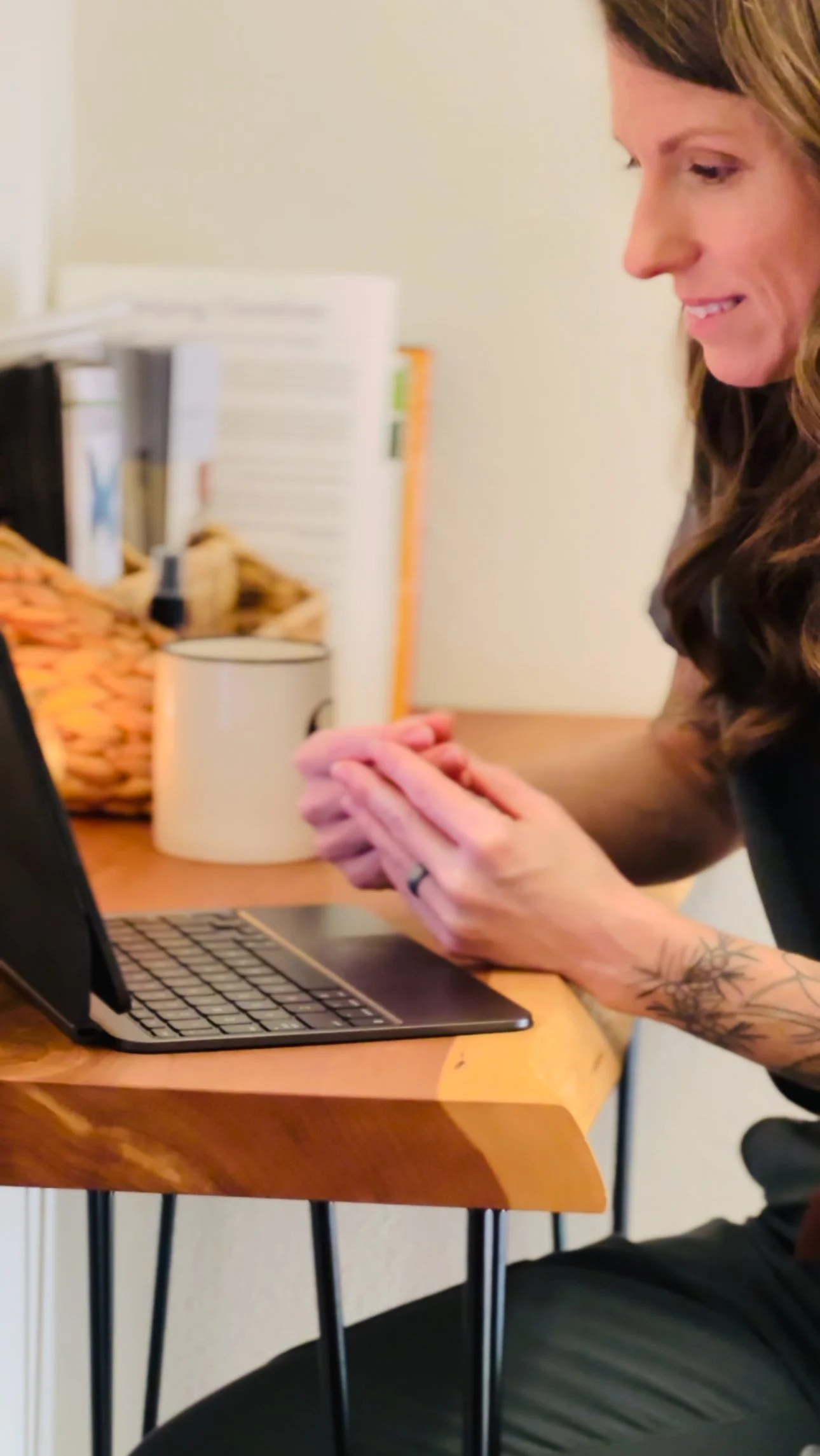 A woman with long wavy hair and tattoos on her arm is sitting at a wooden desk, looking at her phone. A laptop is open in front of her and a white mug is nearby. In the background, there are shelves with books and other items.