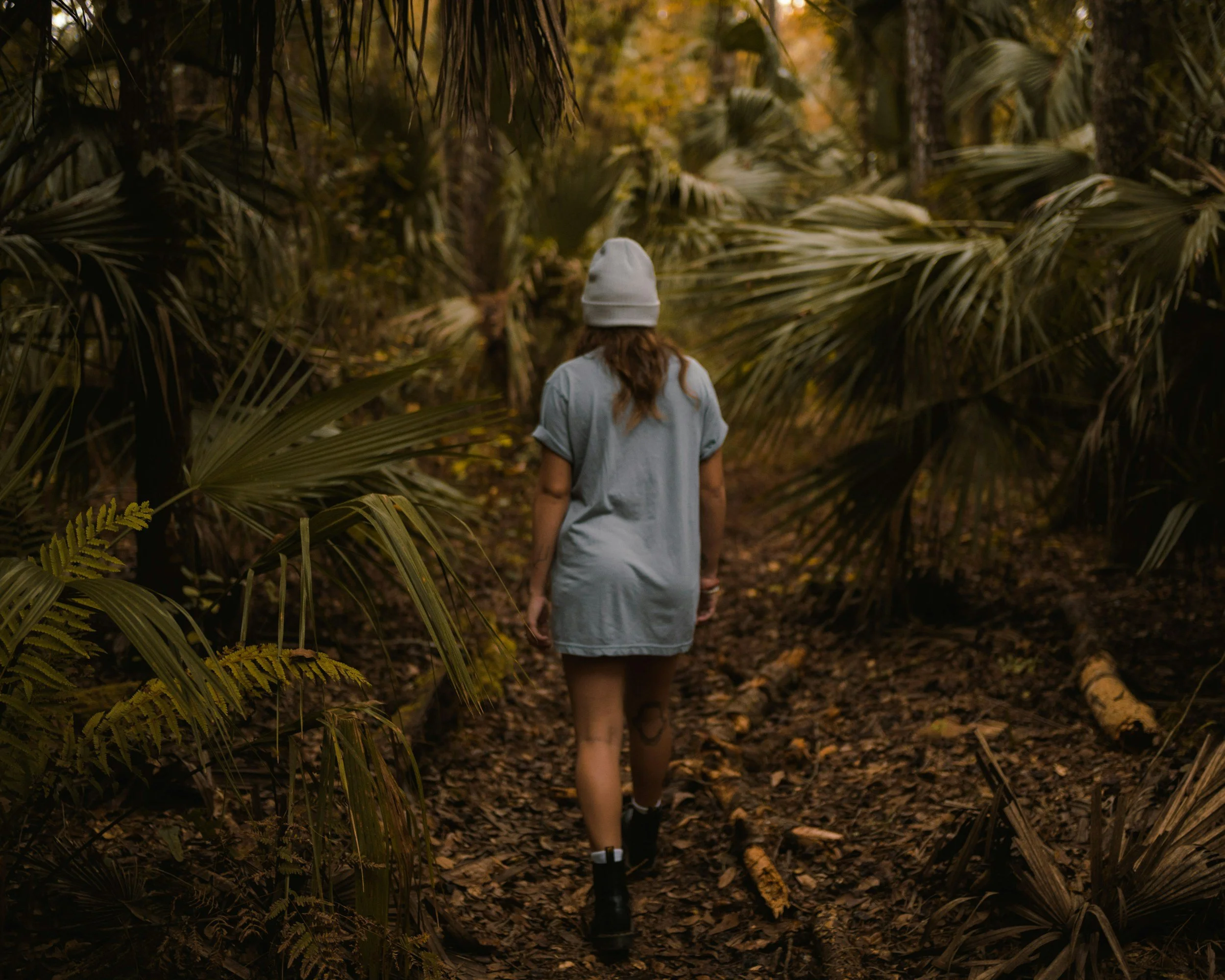 A person with long hair wearing a gray t-shirt, a beanie hat, and black boots walking through a dense tropical forest with large green leaves and a dirt path.