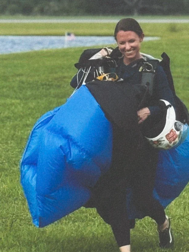 A woman smiling and posing outdoors with skydiving gear, including a helmet and parachute, on a grassy field near a body of water.