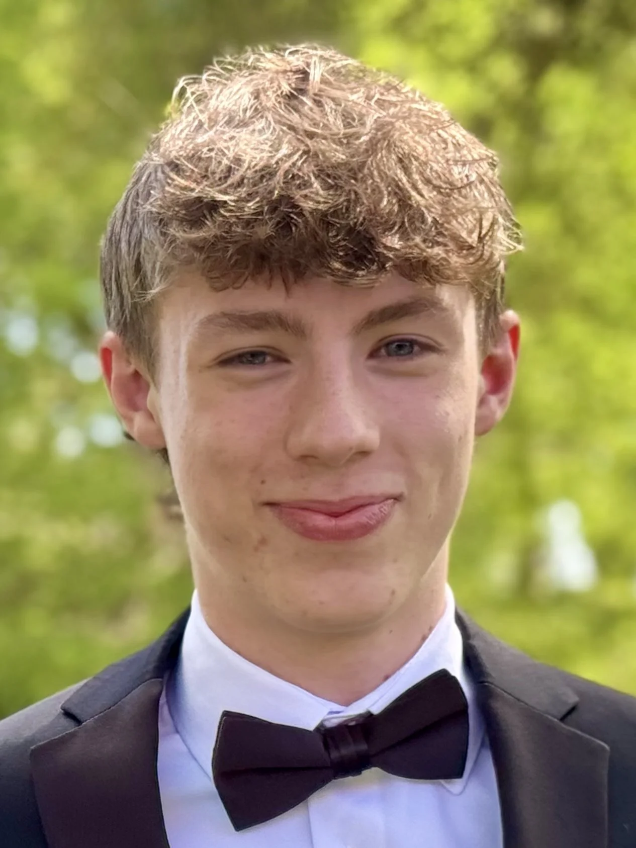 A young man wearing formal attire in an outdoor setting with greenery in the background.