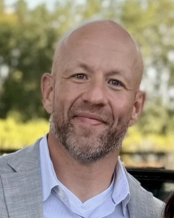 Smiling bald man with a beard wearing a light blazer, outdoors with greenery in the background.
