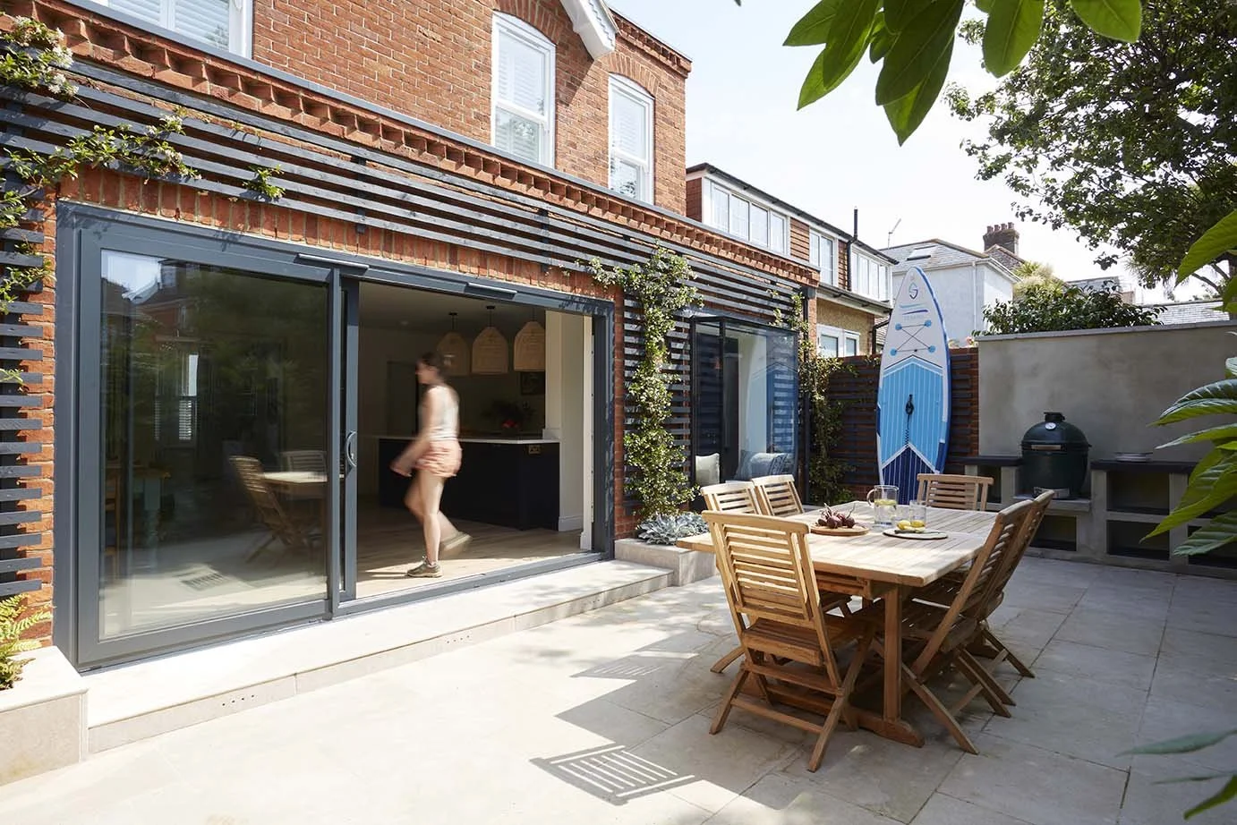 Paved outdoor dining terrace beside a contemporary red-brick house extension, with timber garden furniture, large sliding glass doors and planted boundaries.