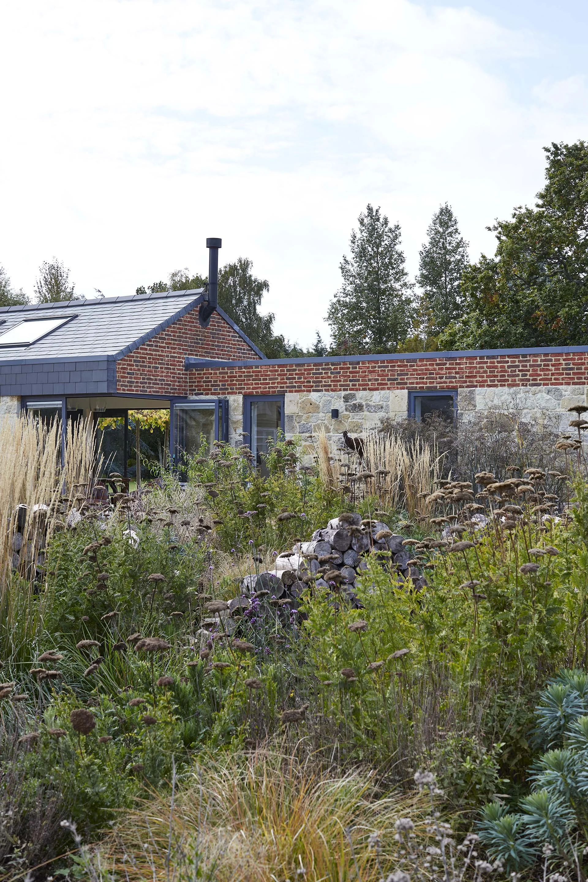 Layered farmhouse garden planting with ornamental grasses and perennials in front of a contemporary stone extension and traditional brick wall.