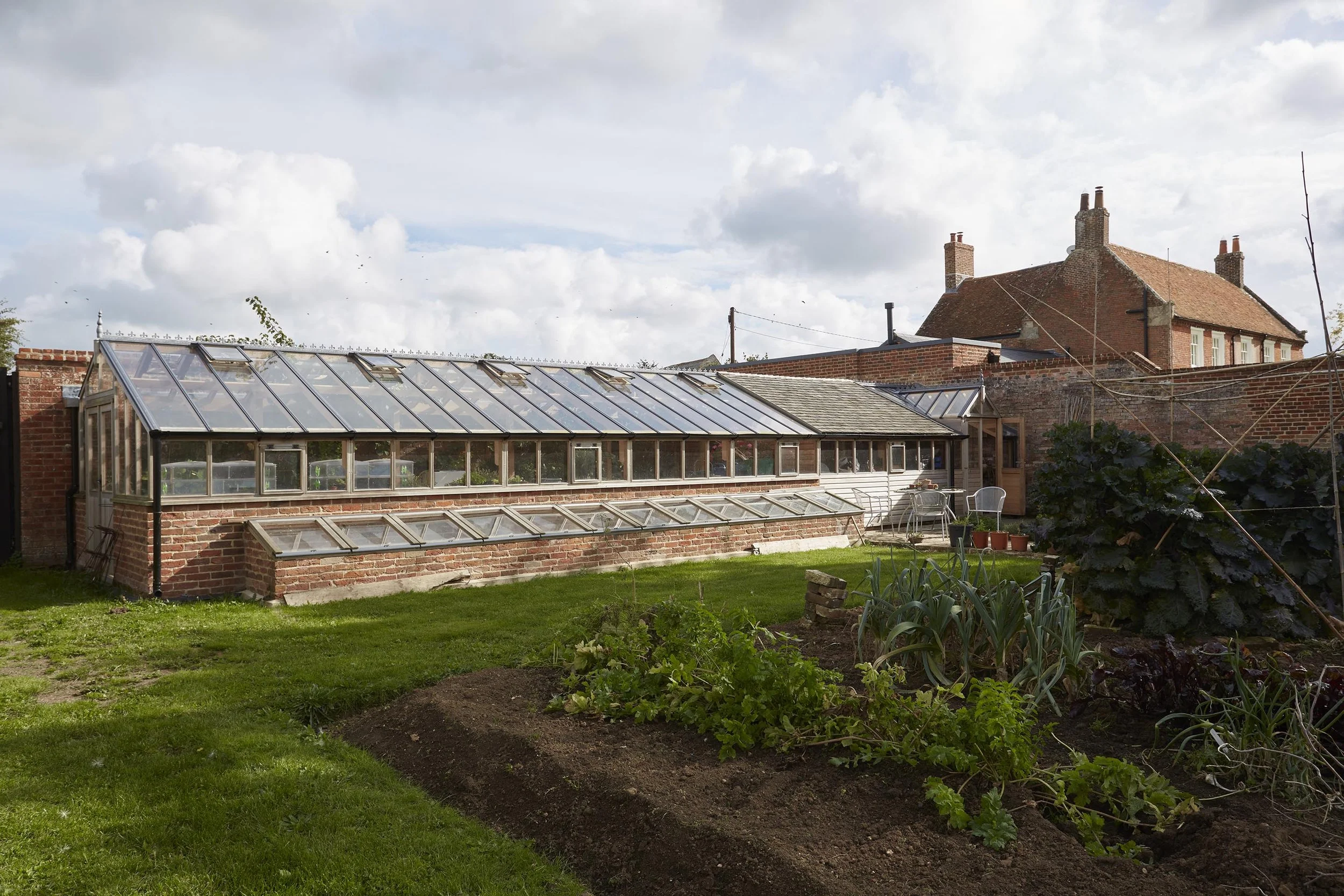 Traditional glass greenhouse with a red-brick base and glazed roof within the historic farmhouse kitchen garden.