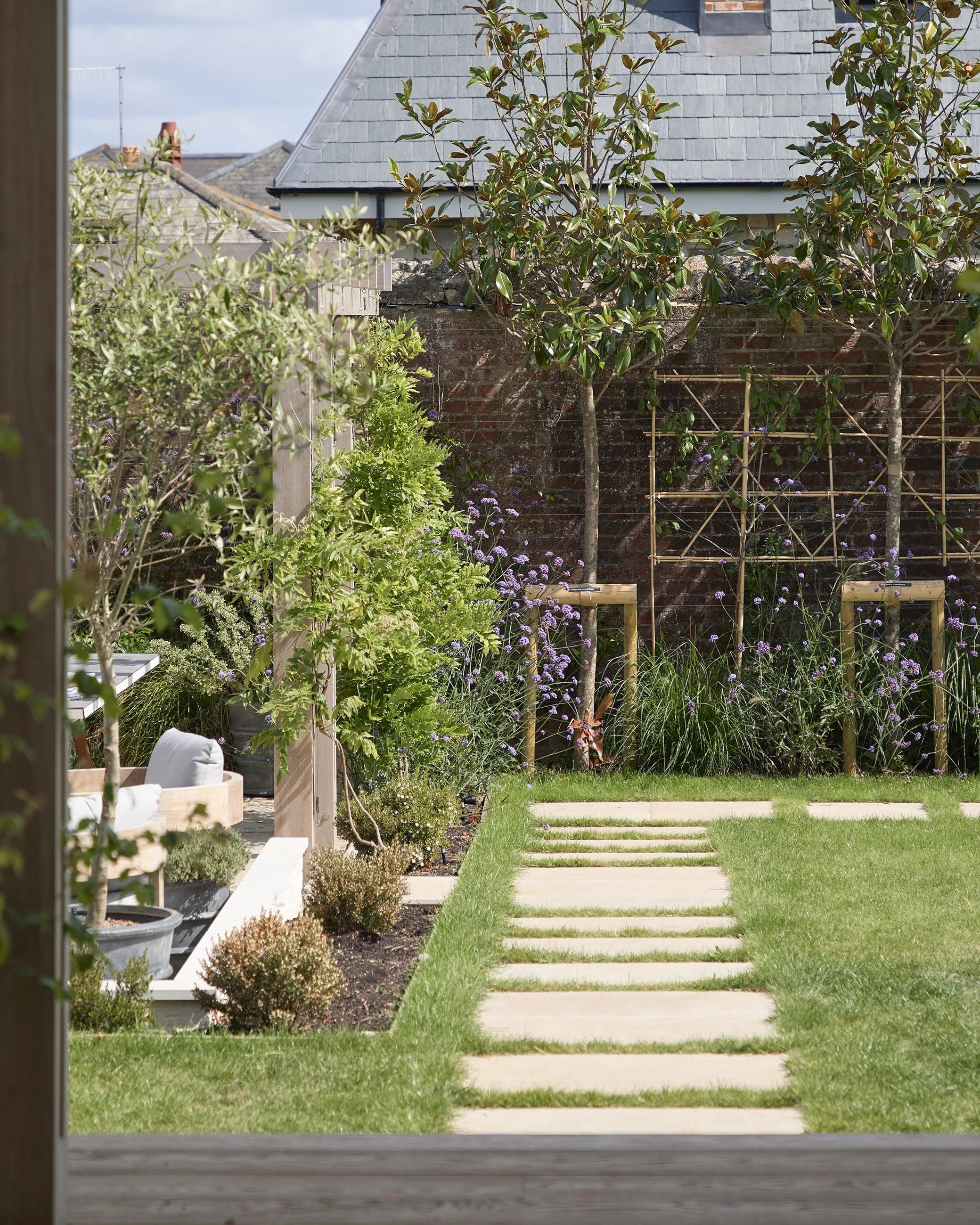 Pale stone stepping path crossing the lawn towards a planted garden border, with young trees, flowering perennials and an existing brick boundary wall.