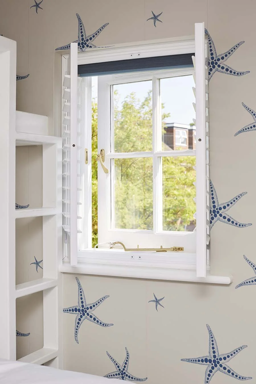White-painted bedroom window and integrated bunk-bed joinery framed by blue starfish wallpaper in a coastal family home.