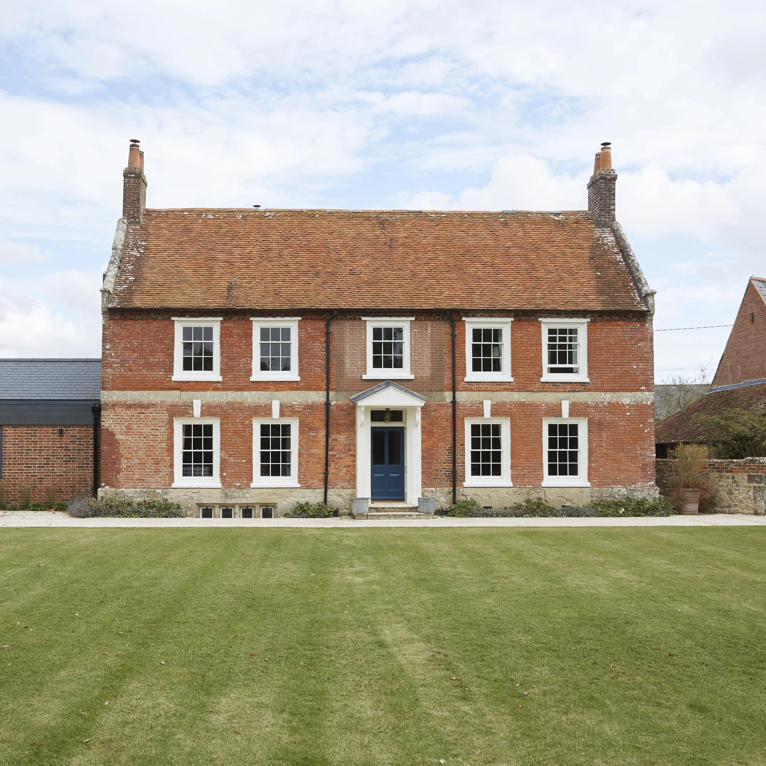Traditional Georgian red-brick farmhouse with symmetrical sash-style windows and tiled roof, alongside a contemporary single-storey extension.