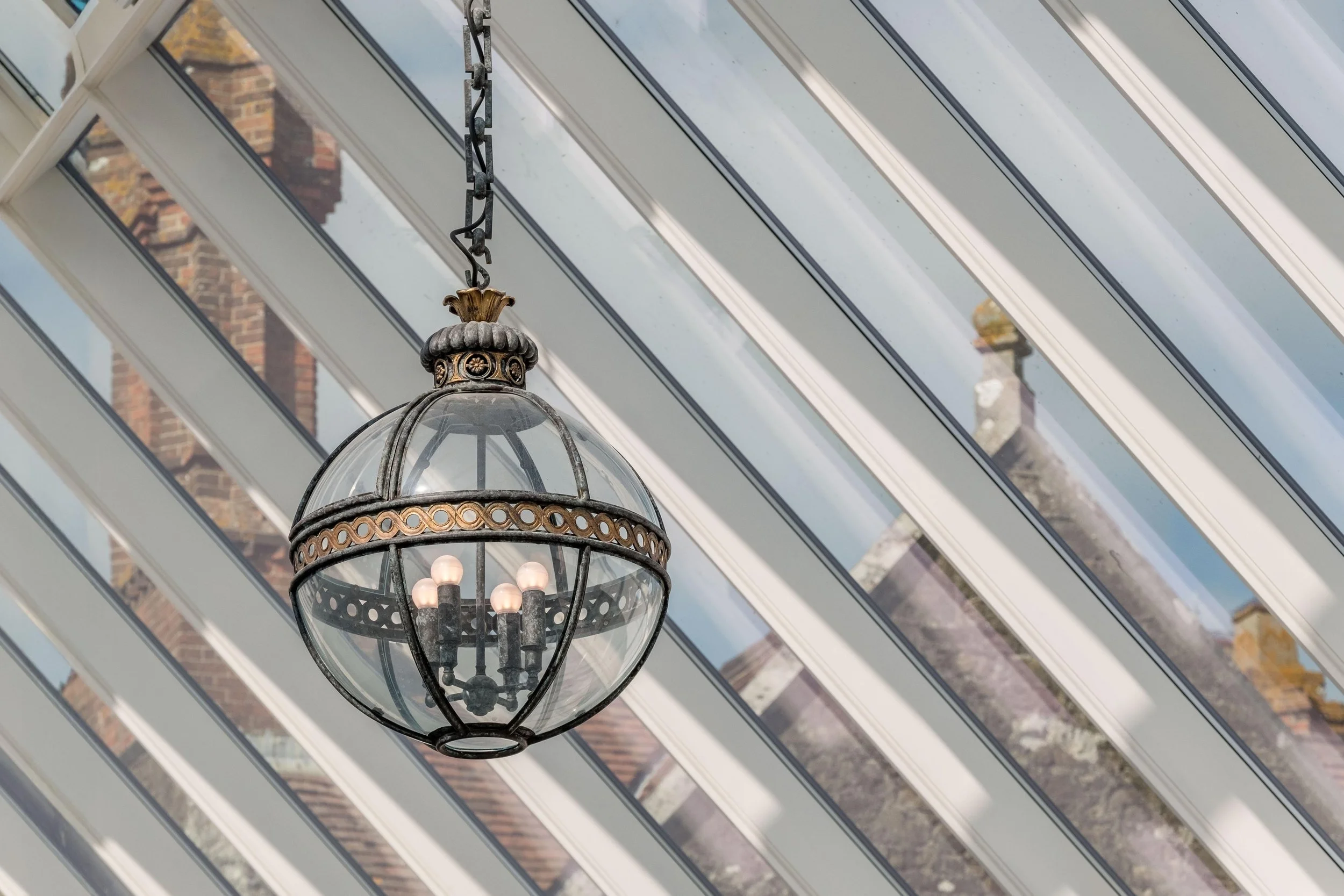 Decorative hanging lantern beneath the glazed roof of the Barton Manor pool enclosure, with historic brick chimneys visible beyond.