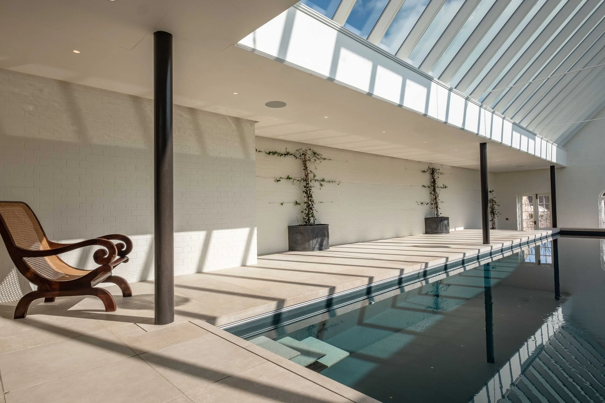 View along the Barton Manor indoor swimming pool, showing the glazed roof, slender dark columns and sunlit poolside.