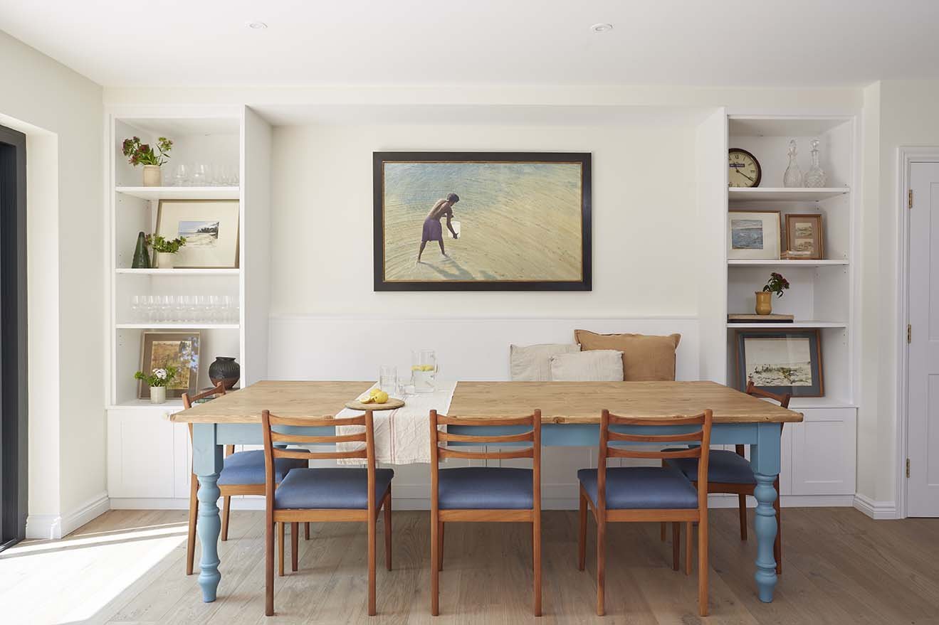 Bright dining room with a pale blue timber dining table, wooden chairs, bespoke white shelving and coastal artwork.