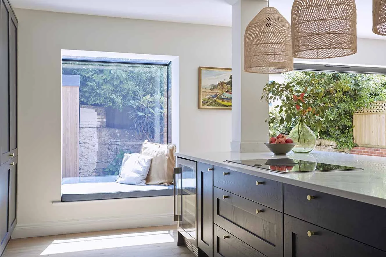 Contemporary kitchen with dark blue cabinetry, pale worktops, woven pendant lights and a large projecting window seat overlooking the garden.