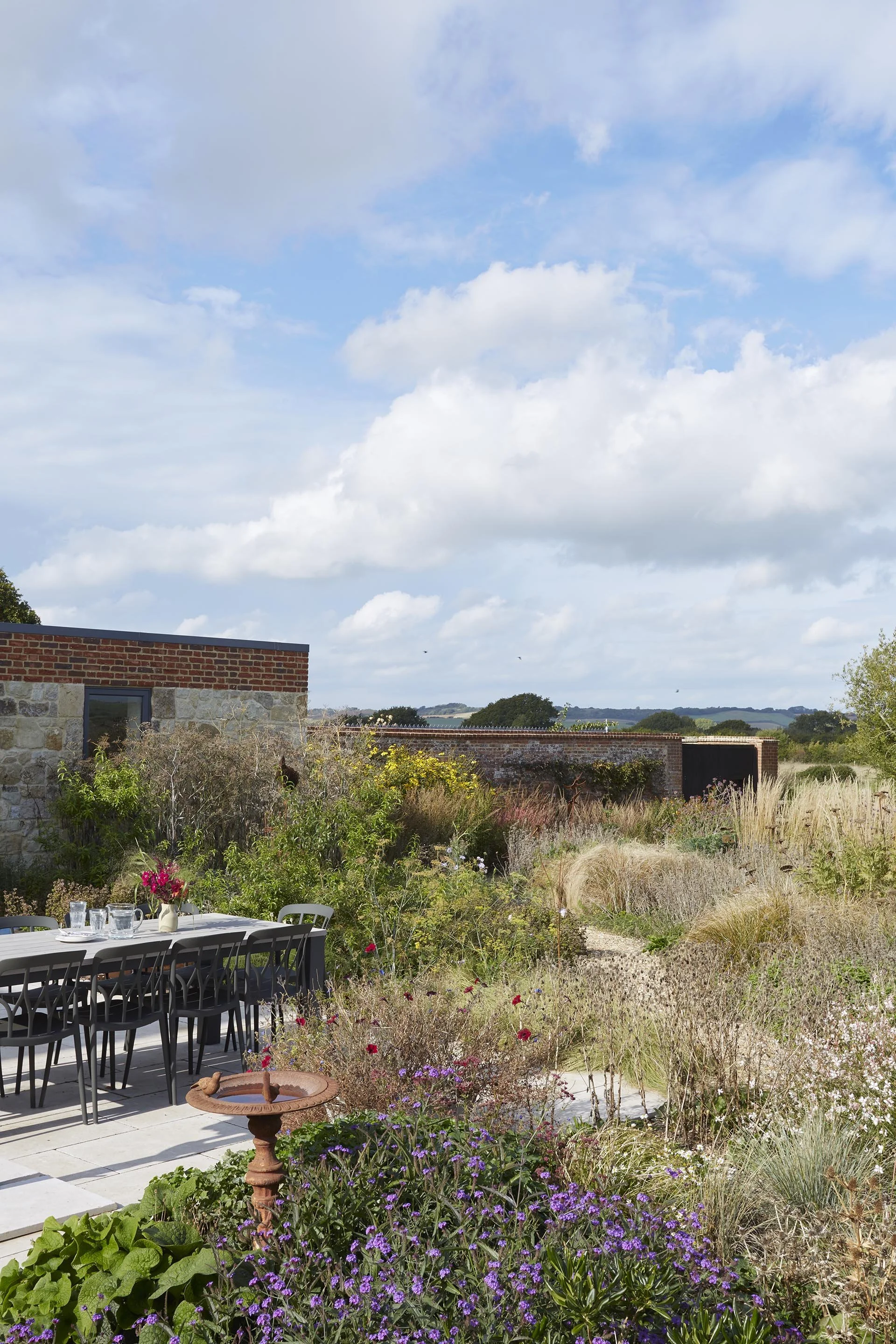 Outdoor dining terrace surrounded by ornamental grasses, purple flowering plants and mixed perennial planting in a rural farmhouse garden.