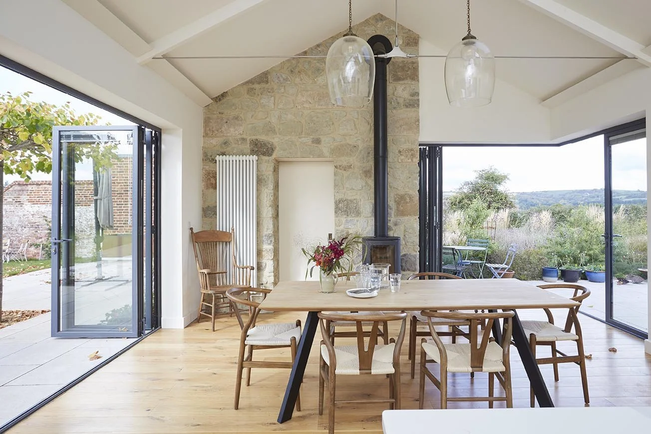 Farmhouse dining room with vaulted ceiling, exposed stone fireplace, timber dining table and large glazed doors opening onto the garden.