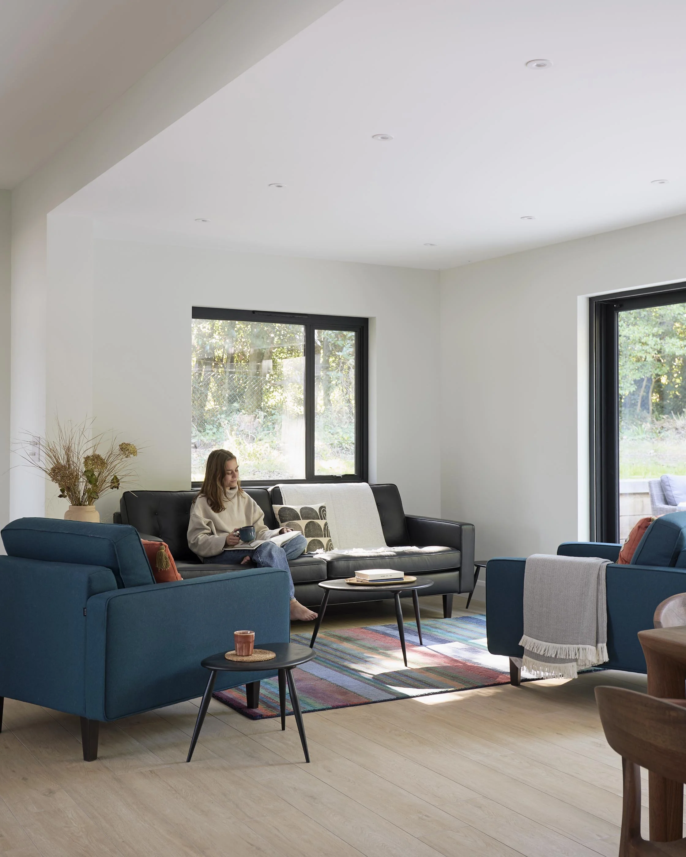 Contemporary living room with blue armchairs, a dark sofa and colourful rug, framed by large black-framed windows overlooking greenery.