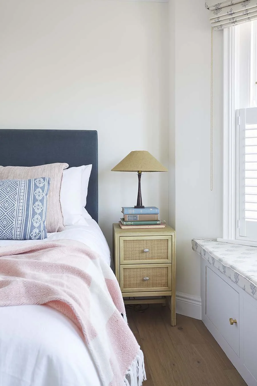 Calm coastal bedroom with a blue upholstered headboard, soft pink bedding, a natural woven bedside table and warm bedside lighting.