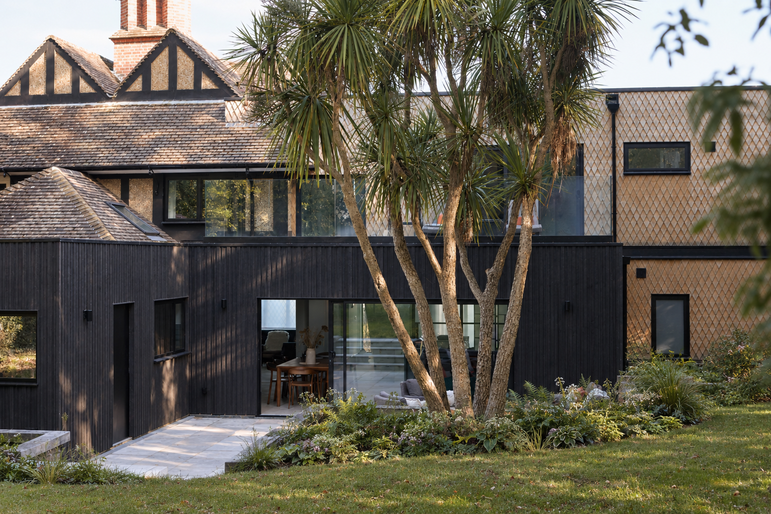 Contemporary oak-shingle and dark timber extension set within a landscaped garden with mature trees and stone paving.