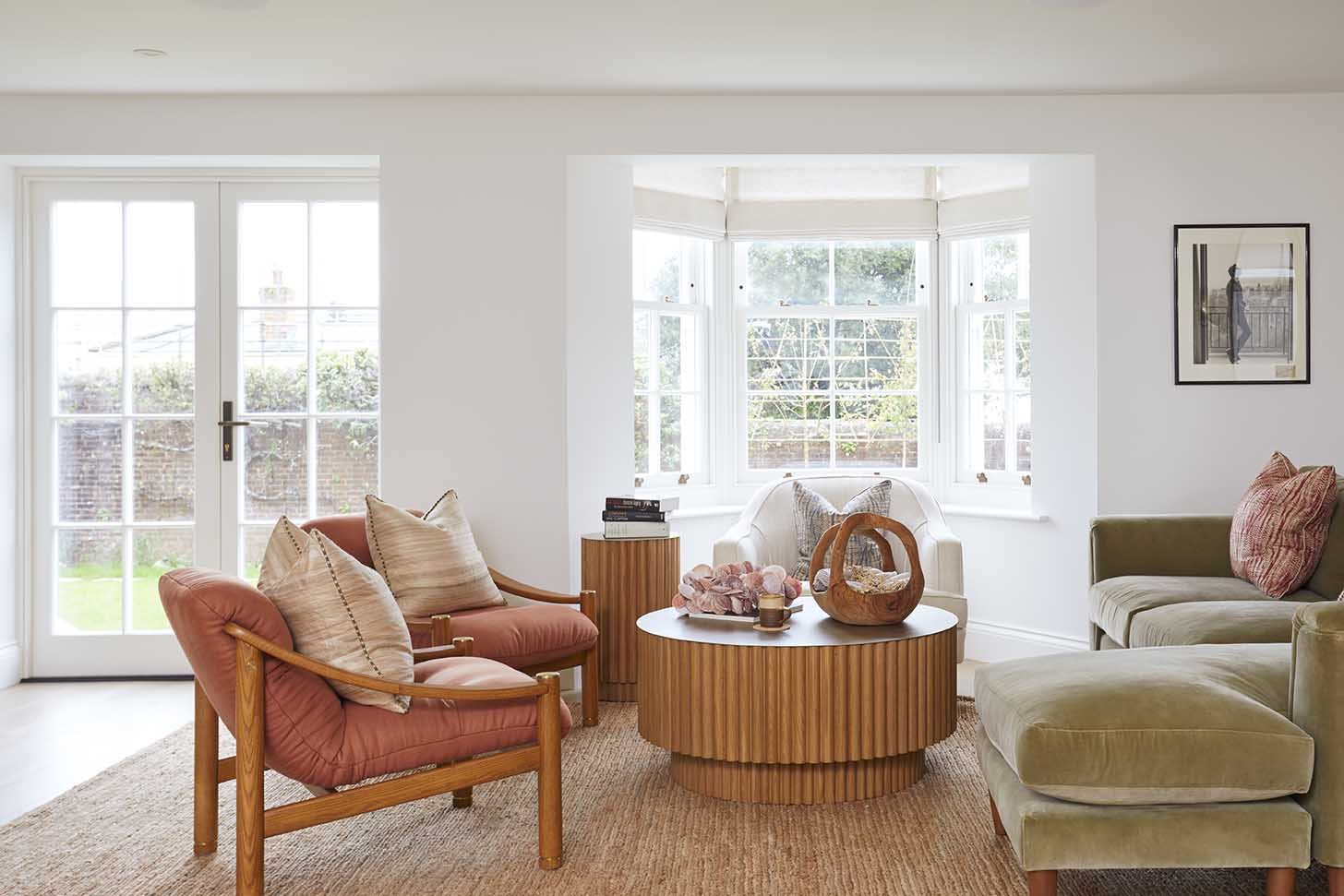 Contemporary living room with salmon-pink armchairs, a sage-green sofa and a round fluted timber coffee table, creating a relaxed coastal interior.