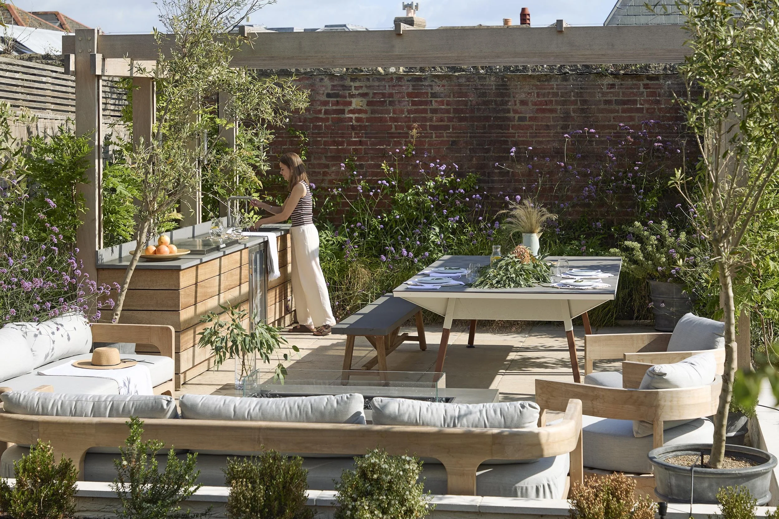 Outdoor kitchen and dining terrace beneath a timber pergola, with built-in timber cabinetry, garden seating and layered planting for outdoor entertaining.
