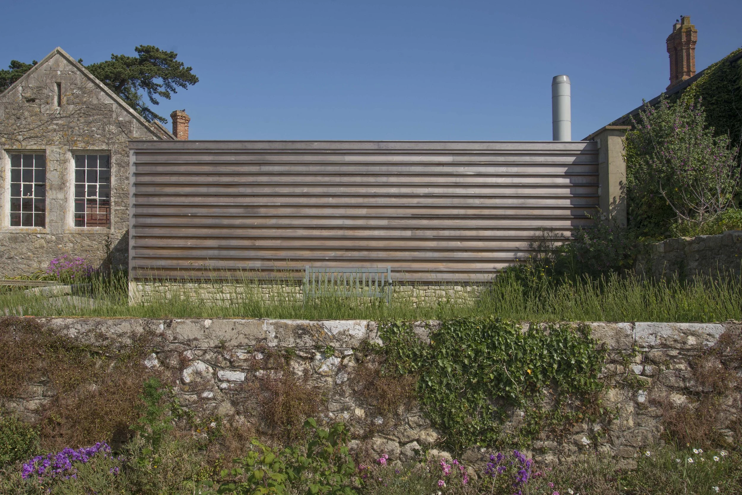 Contemporary horizontal timber-clad structure at Barton Manor Estate, set against traditional stonework and established garden planting.