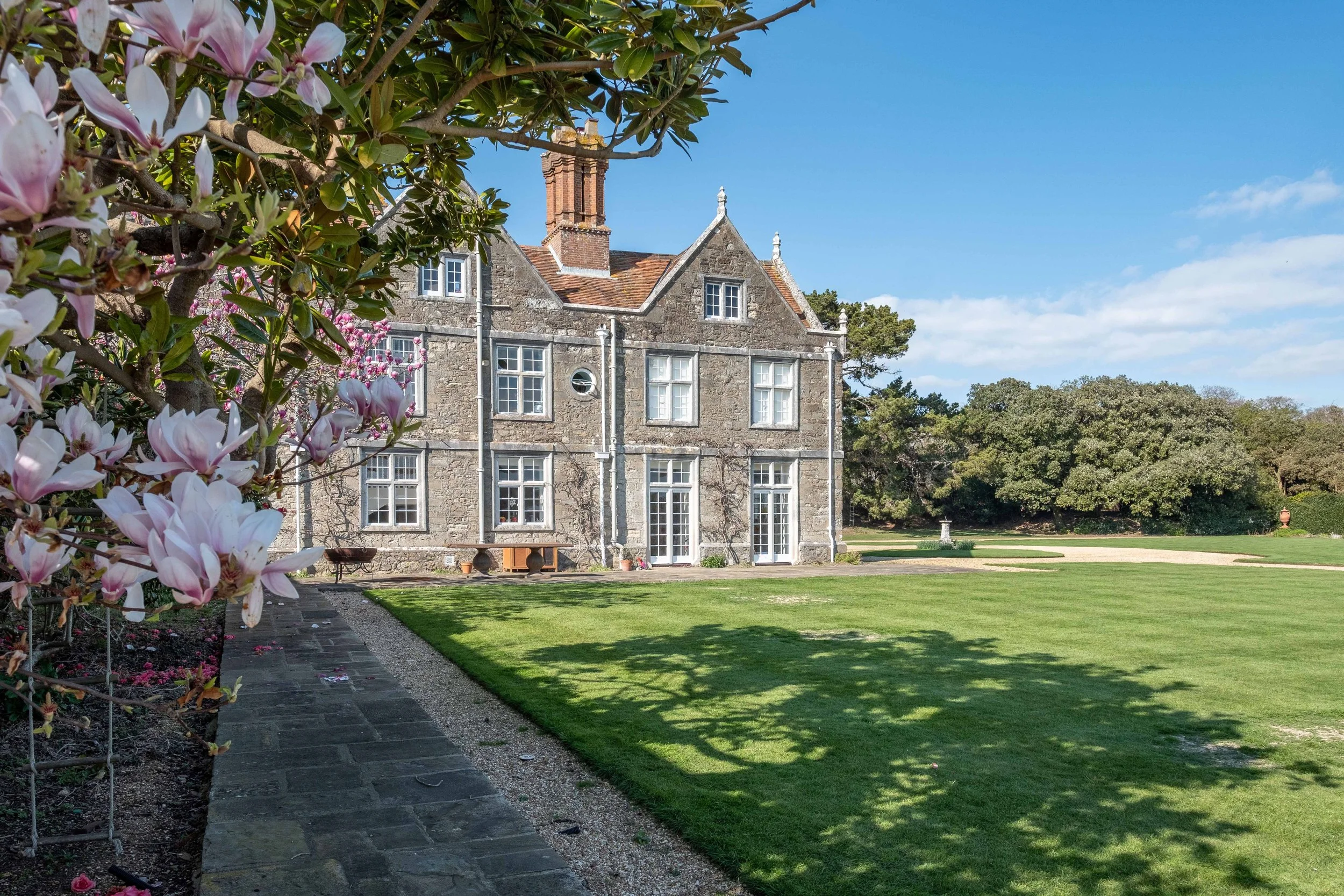 Historic Jacobean manor house at Barton Manor Estate, viewed across the lawn and garden path with flowering magnolia in the foreground.