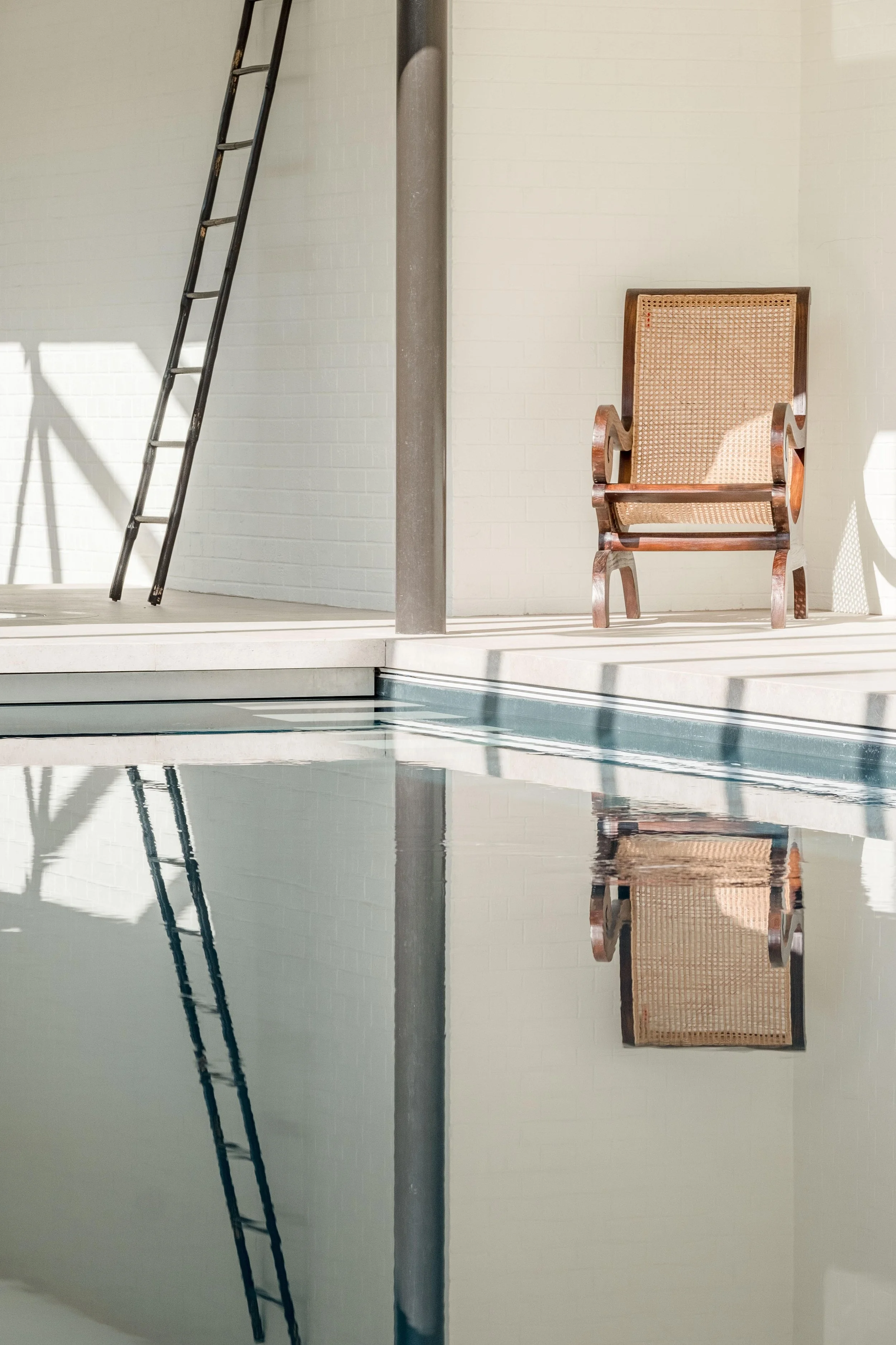 Detail of the indoor pool at Barton Manor Estate, with a cane chair reflected in the water beside the white brick wall.