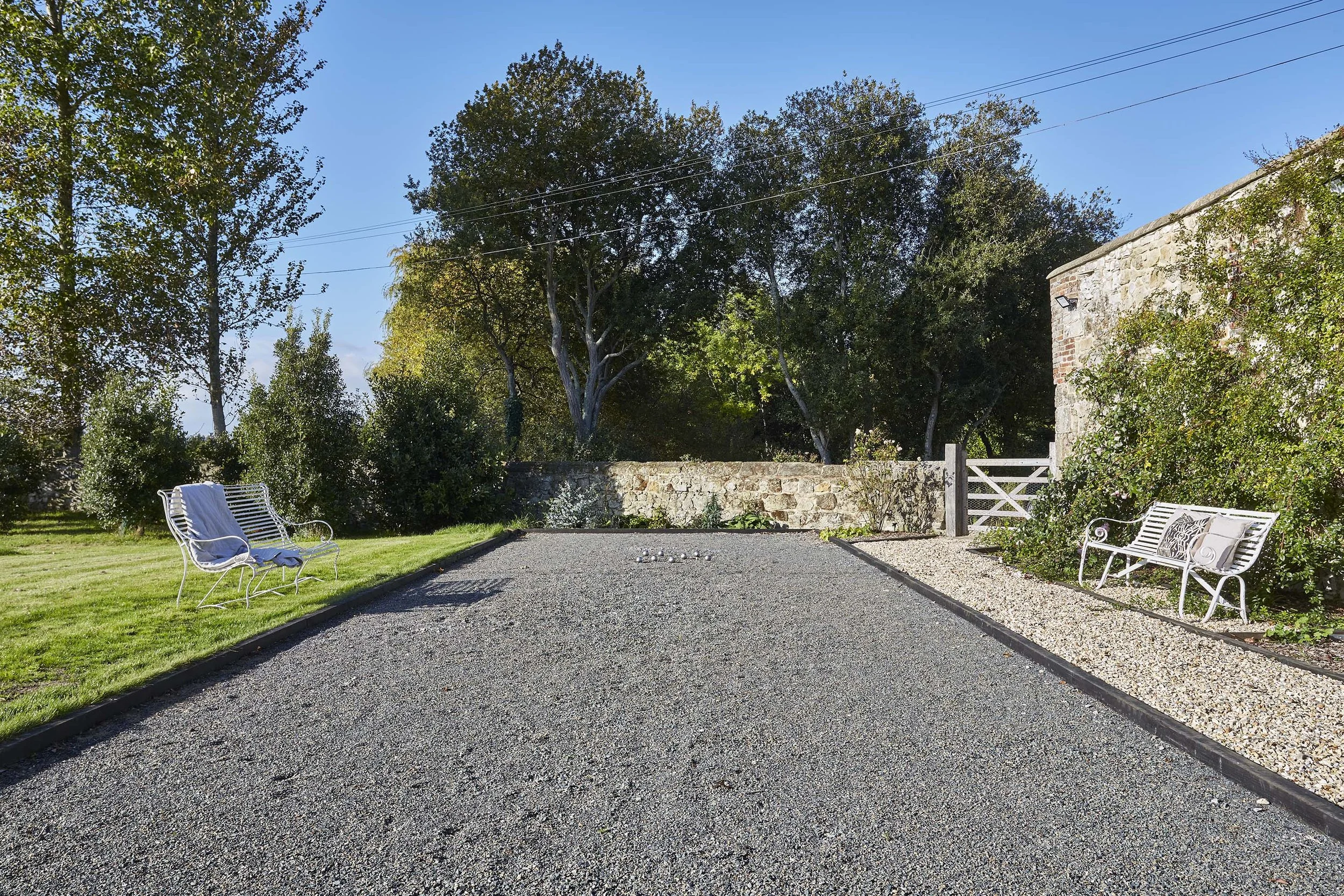 Gravel courtyard beside a traditional stone outbuilding, with white metal garden benches, planted borders and mature trees.