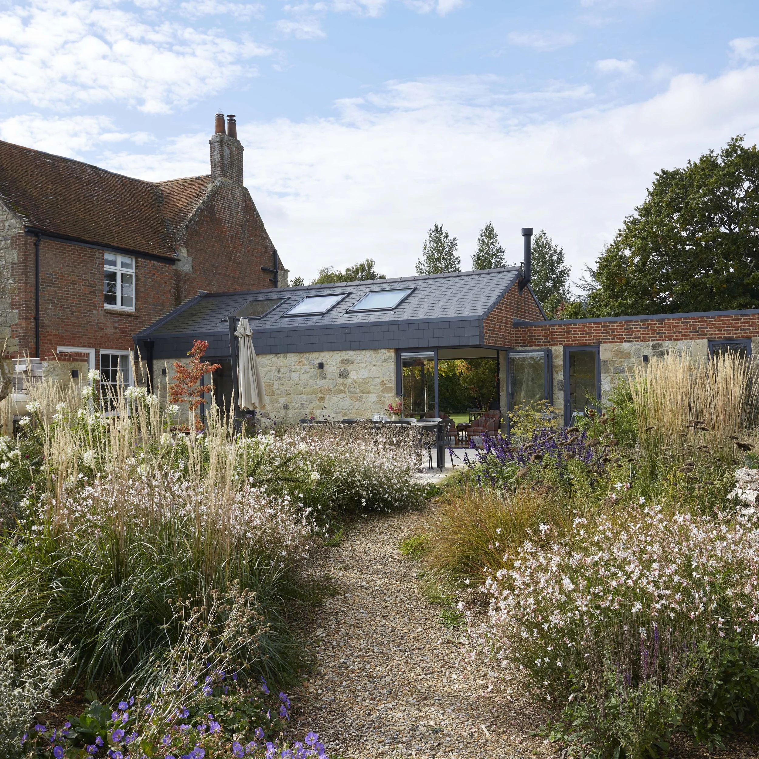 Contemporary stone and brick extension with slate rooflights, set within a landscaped farmhouse garden with ornamental grasses and gravel paths.