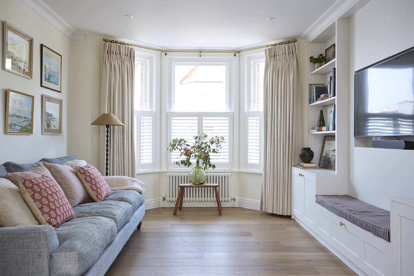 Coastal living room with a blue upholstered sofa, bay window with white shutters and bespoke fitted shelving and media storage.