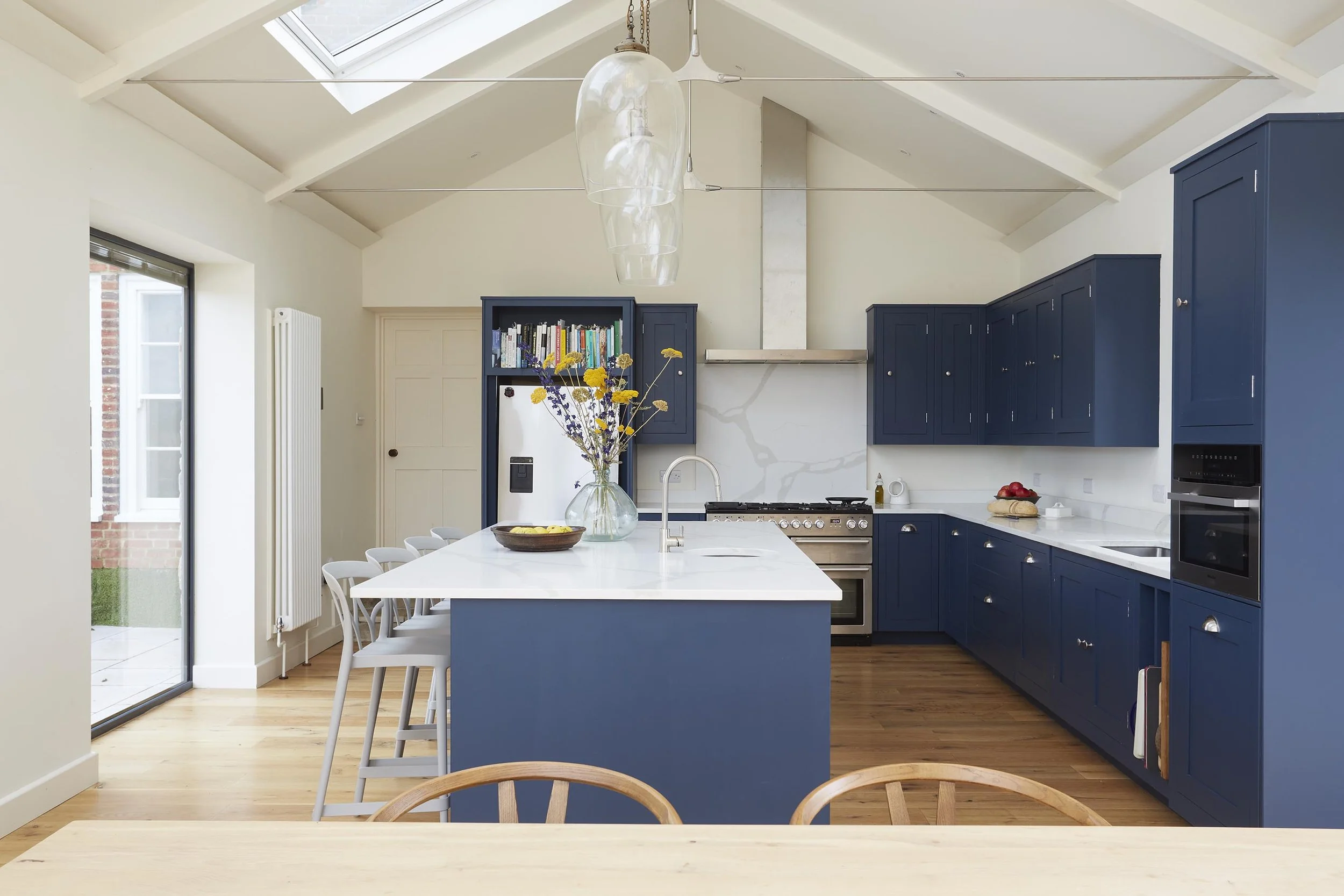 Bespoke navy-blue farmhouse kitchen with central white island, timber flooring, vaulted ceiling and rooflight above the cooking area.