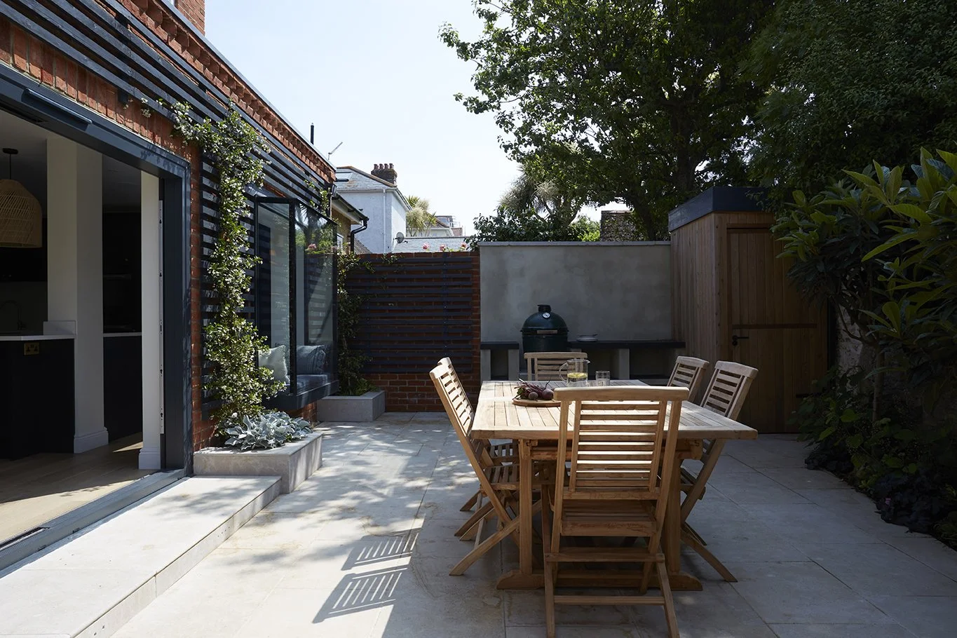 Paved outdoor dining terrace beside a contemporary red-brick house extension, with timber garden furniture, large sliding glass doors and planted boundaries.