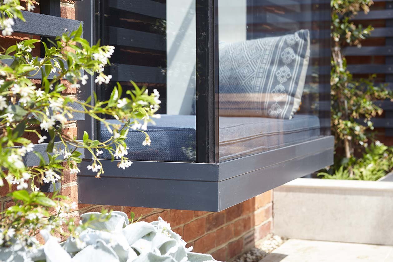 Bespoke projecting window seat with dark metal framing, upholstered cushions and climbing plants against the red-brick extension.