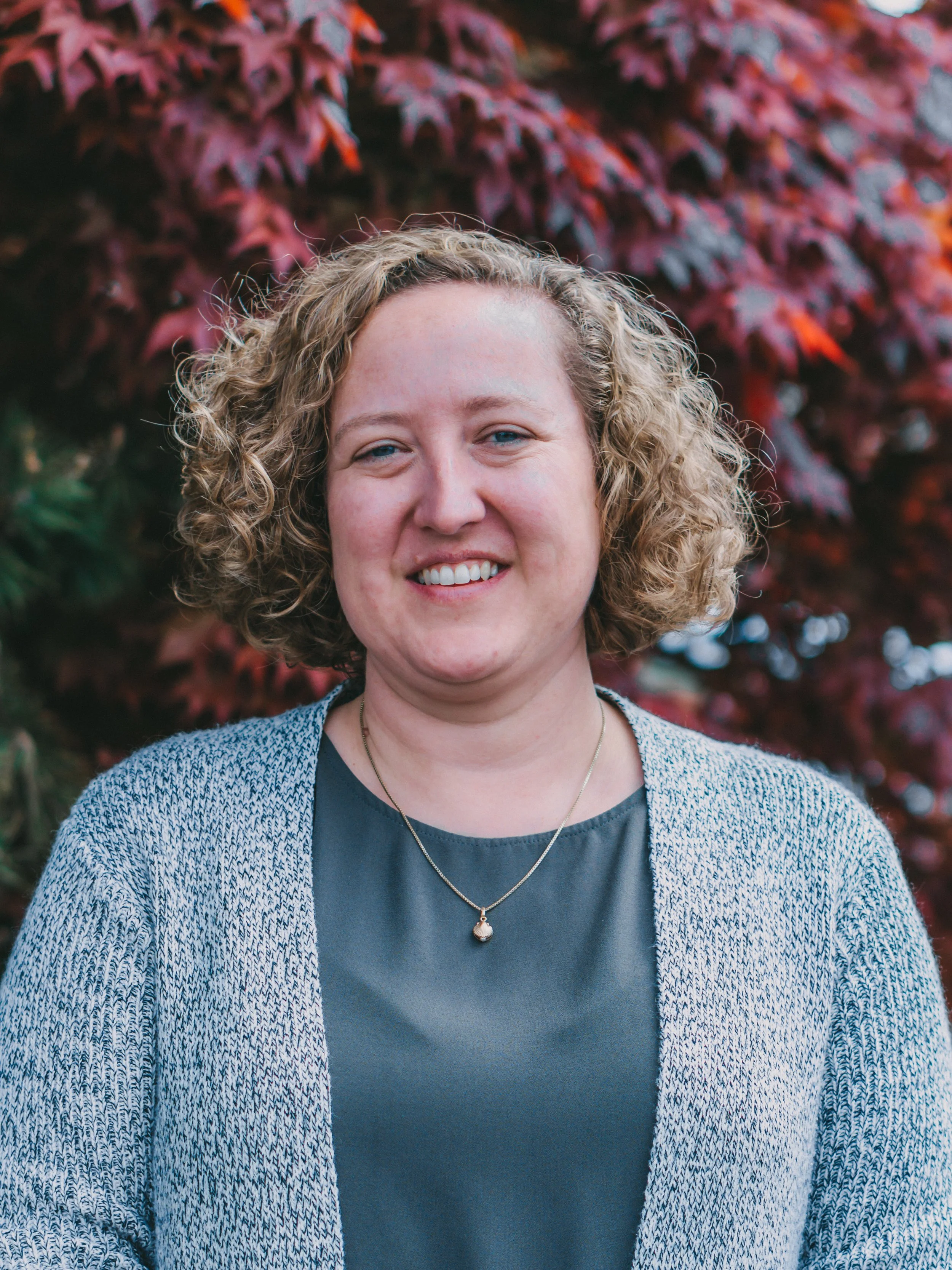 Person with curly hair, wearing blue top and gray cardigan, standing in front of red leaves.
