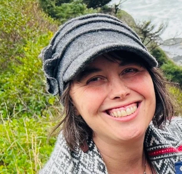 Person wearing a colorful hat and mustard yellow striped blouse, standing outdoors near a pond with autumn foliage.
