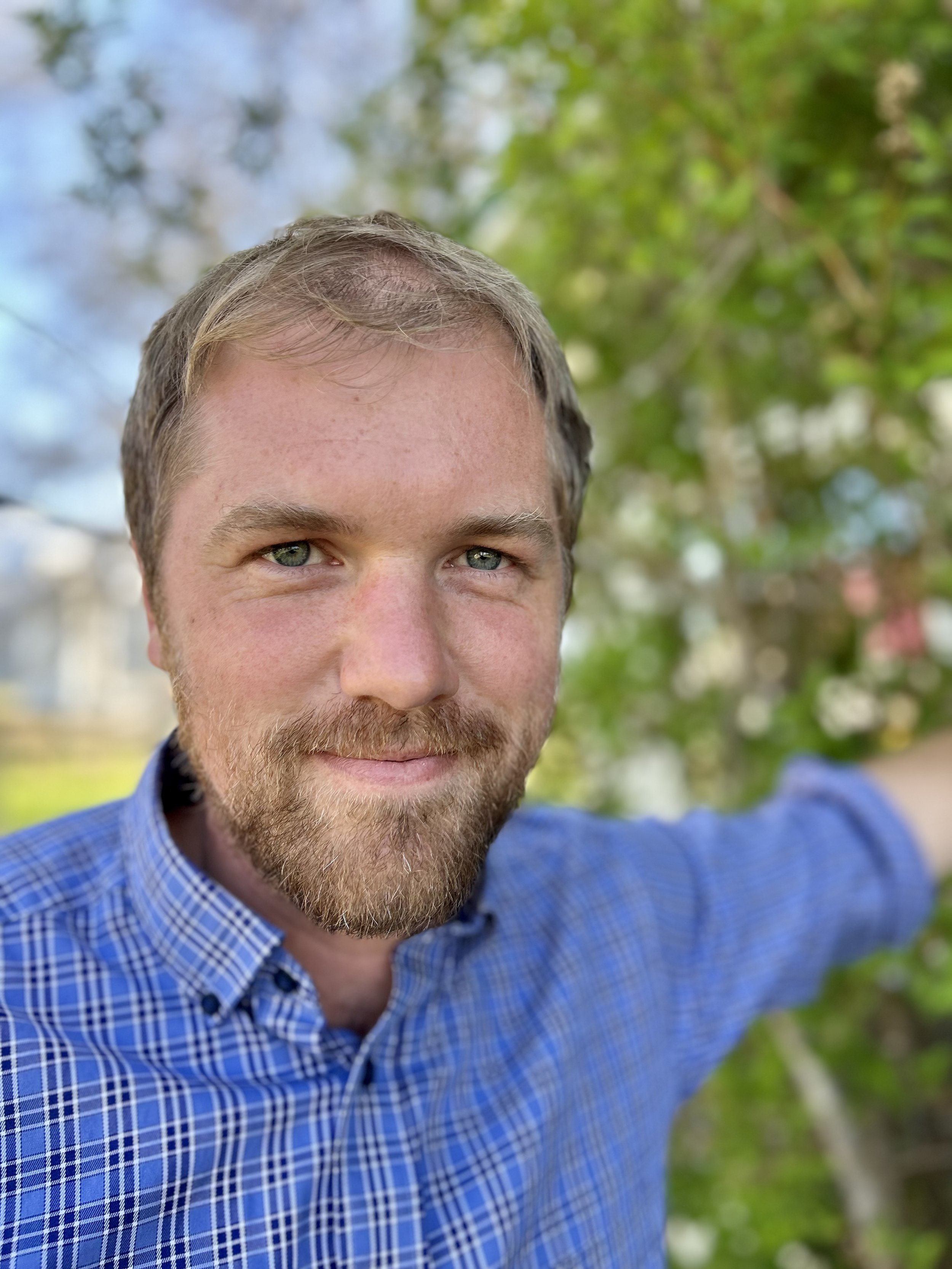 A man with a beard and blue eyes smiling outdoors, wearing a blue checkered shirt, with green leaves and a tree in the background.