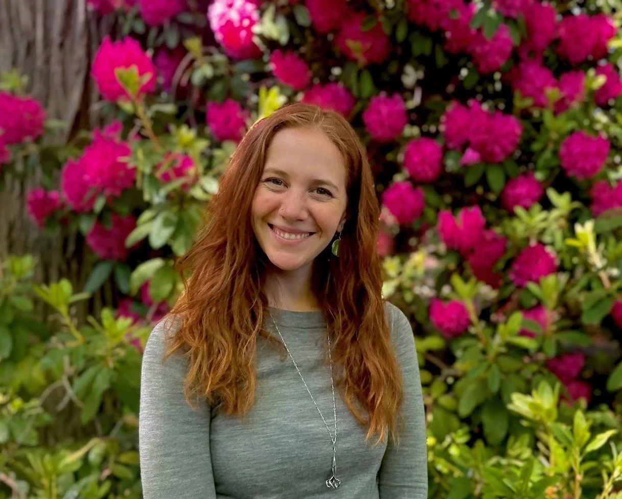 Woman with red hair smiling next to a tree, wearing a green jacket in a forest setting.