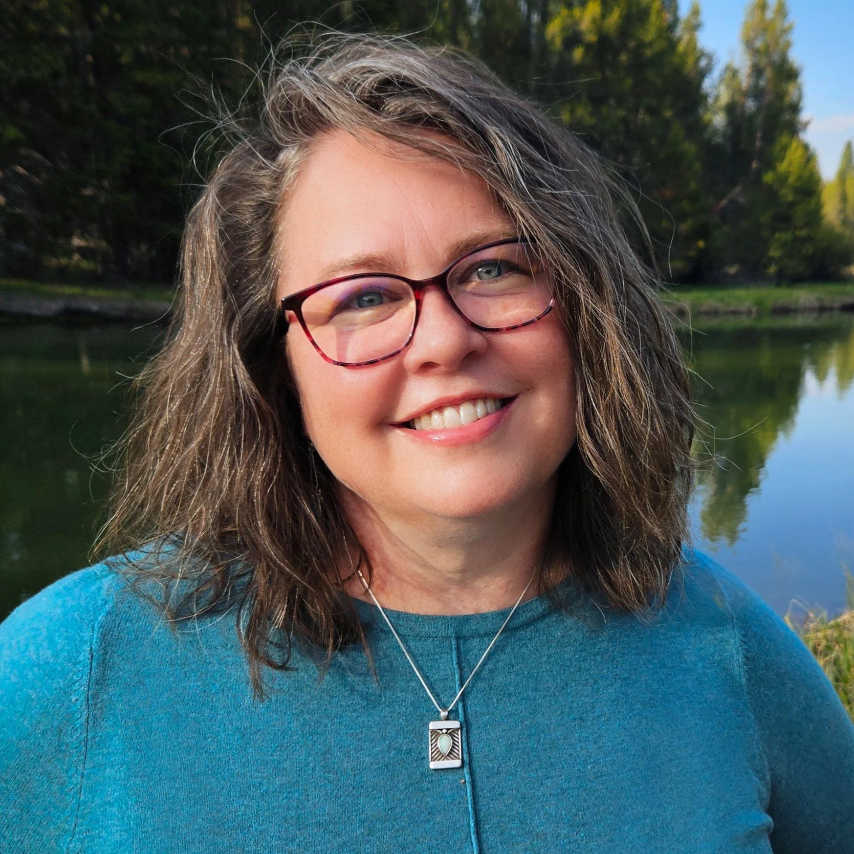 Smiling woman with glasses and wavy hair, wearing a blue sweater, in an outdoor setting with trees and a lake in the background.