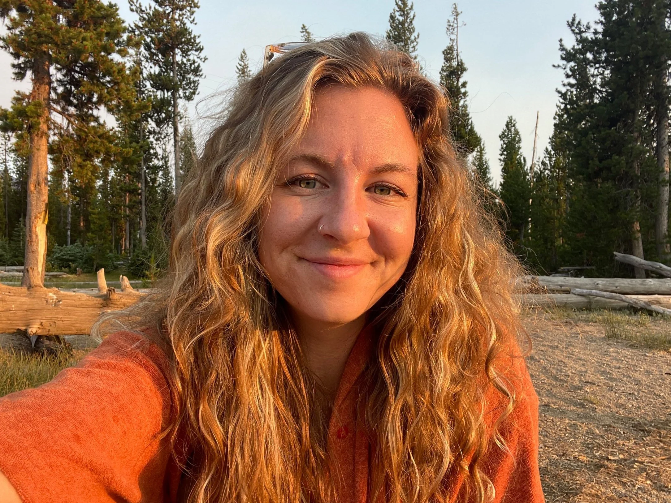 Person with long curly hair smiling outdoors, surrounded by trees and logs, under a clear sky.