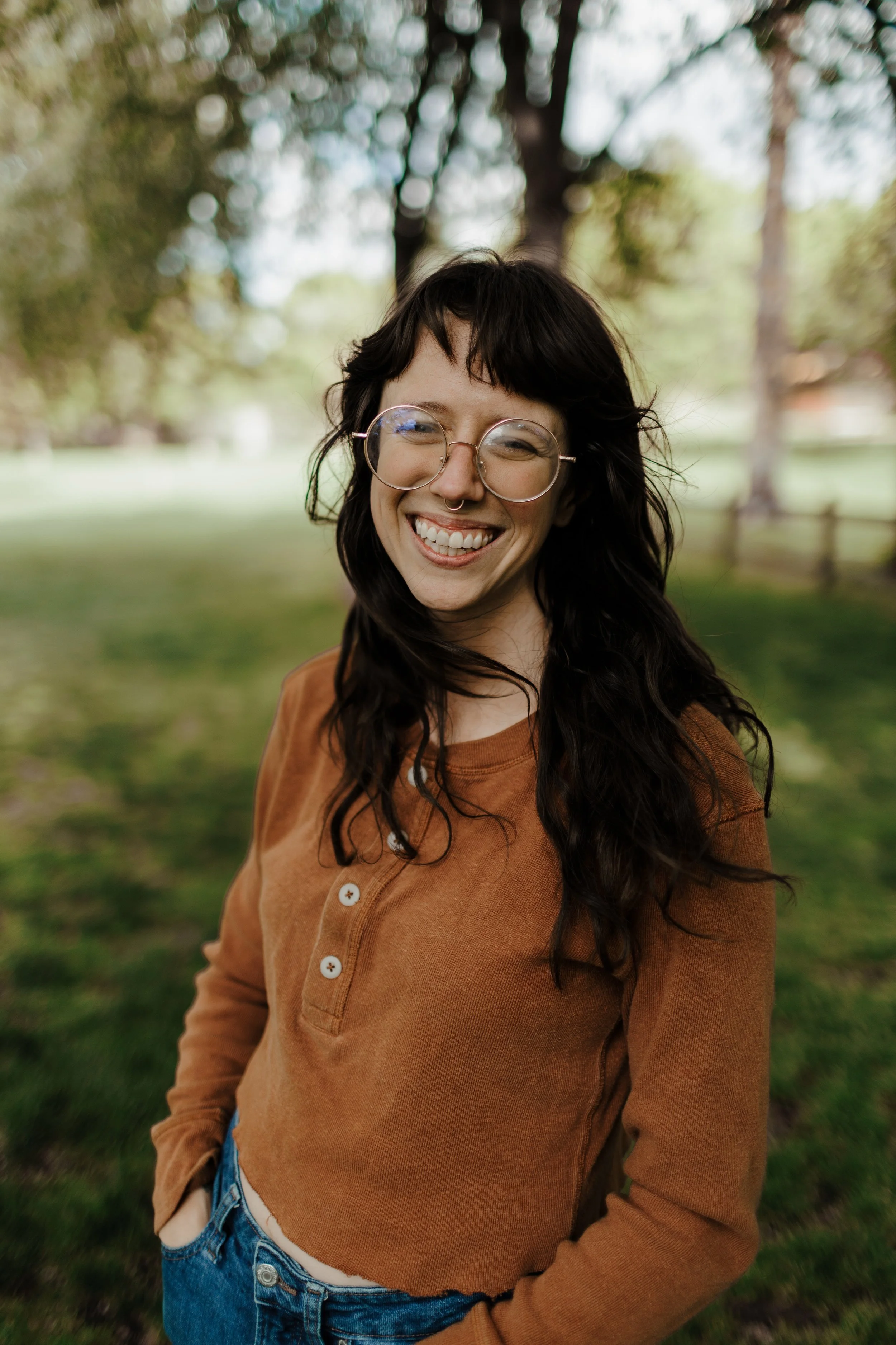Person with long brown hair and glasses smiling outdoors in a park, wearing a brown shirt and blue jeans, with trees in the background.