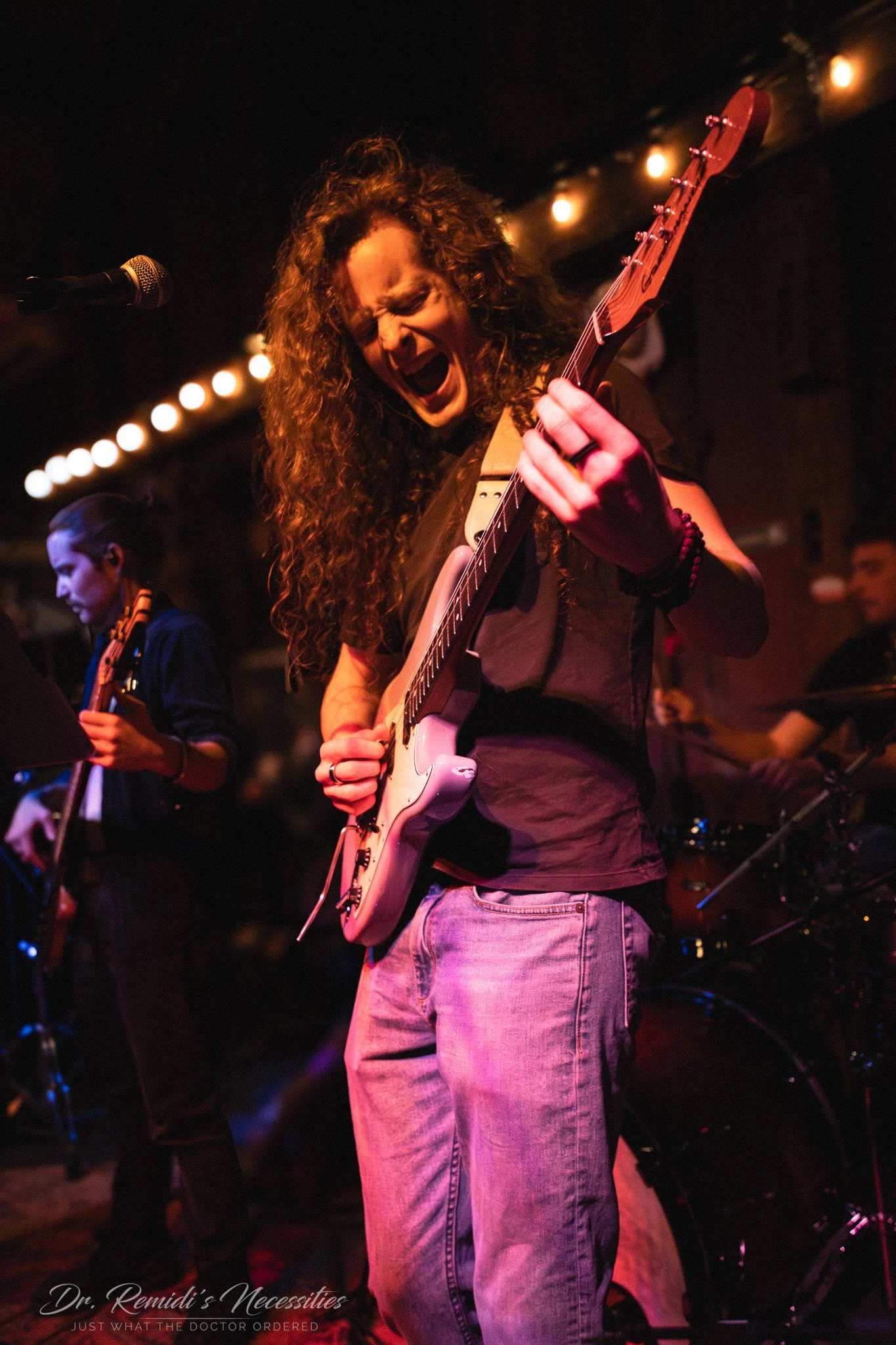 A woman with curly hair passionately playing an electric guitar on stage, with a band performing in the background under warm stage lighting.